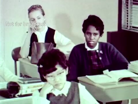 The still features three students in a classroom setting. Two girls are sitting at desks, one appears bored while the other looks contemplative. The third girl has glasses and a focused expression, seemingly engaged with her work. The film emphasizes the integration of IBM computer technology in educational settings, showcasing its application in handling grades and course enrollment during the 1960s. The educational context highlights the potential benefits of technology in streamlining administrative processes.