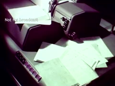 The still shows a vintage IBM computer setup from the 1963 film 'IBM - The Information Center.' It features two large, boxy machines, likely printers or card readers, with sheets of paper scattered around them. The documents appear to involve data entry, possibly related to academic records such as grades or enrollment. The overall ambiance reflects the technological capabilities of IBM at the time, highlighting the use of punch cards in educational administration.