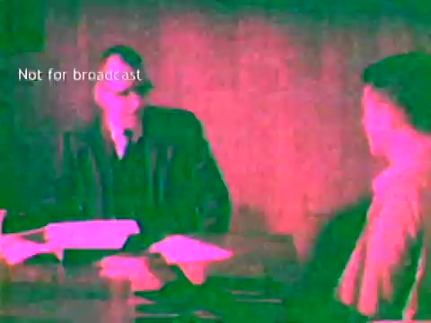 The still shows two men engaged in a conversation at a table, likely discussing educational processes related to IBM's technology. One man is dressed in a suit and appears to be explaining something, while the other, dressed more casually, listens attentively. The setting has a muted color palette typical of 1960s film, and there is a watermark stating 'Not for broadcast,' indicating its intended non-commercial use. The focus is on the use of IBM's computing technology, such as punch cards, in educational contexts.