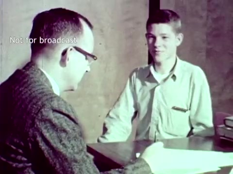 The 16mm still from 'IBM - The Information Center' (1963) features a conversation between an adult man, likely an instructor or administrator, and a young student. The setting suggests an educational environment, with a desk and books visible. The adult, wearing glasses and a patterned jacket, appears to be discussing academic matters with the student, who is looking engaged and attentive. This scene underscores the application of IBM's computer technology in managing educational tasks, such as grades and enrollment, using tools like punch cards.