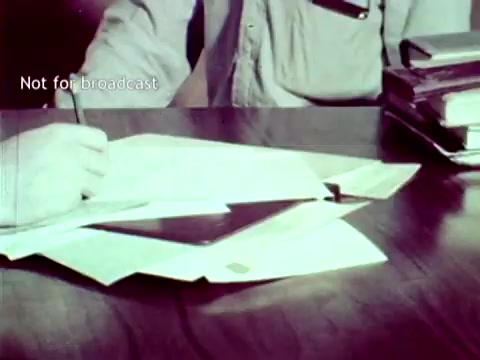 The 16mm still from 'IBM - The Information Center (1963)' depicts a person's hand writing on a sheet of paper. The scene emphasizes the administrative tasks associated with education, showcasing documents related to grades and course enrollment spread across a wooden table. The image highlights the integration of IBM technology, such as punch cards, in managing educational processes during that era. The 'Not for broadcast' label suggests it was intended for internal or promotional use rather than public airing.