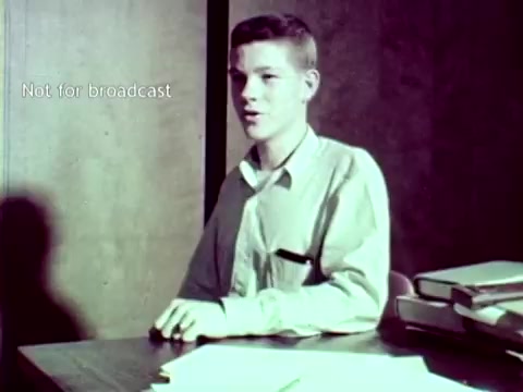 The still from 'IBM - The Information Center (1963)' features a young male student sitting at a desk. He appears to be engaged in a discussion or presentation related to educational technology. The desk is cluttered with papers, suggesting an academic context. The colors are muted, typical of film from that era, and there's a note that indicates the image is not for broadcast. The scene likely illustrates how IBM's technology, such as punch cards, was integrated into managing educational tasks, including grading and course enrollment.
