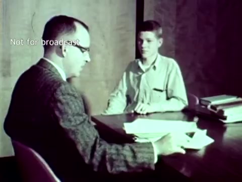 The still depicts a scene from 'IBM - The Information Center' (1963), showing two individuals seated at a table. One is a man in a suit and glasses, engaging in conversation with a younger person dressed casually. Papers and materials are spread across the table, likely related to educational administration, reflecting how IBM's technology, such as punch cards, was utilized for managing grades and course enrollment in educational settings. The image conveys a sense of collaboration and innovation in educational technology.