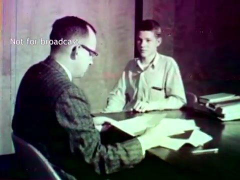 The still features two individuals at a table, one adult and one adolescent. The adult, wearing glasses and a patterned suit, appears to be engaged in a discussion with the younger person, who looks attentive. Papers are spread on the table, which may include documents related to educational records. The setting suggests an academic context, likely a meeting to discuss course enrollment or grades, reflecting the use of IBM technology in educational administration during the 1960s. The overall tone is informative, highlighting the interaction and reliance on technology of the era.