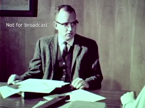 The still from 'IBM - The Information Center (1963)' features a man in a tweed jacket seated at a desk, engaged in conversation. The setting appears to be an office with wooden paneling. Documents and punch cards are visible on the desk, illustrating the use of IBM technology in managing educational tasks like grades and course enrollment. The text 'Not for broadcast' indicates the image is intended for internal or archival purposes rather than public viewing.