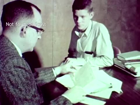 The still from 'IBM - The Information Center (1963)' features two individuals seated at a table, engaged in discussion. One person, wearing glasses and a suit, appears to be reviewing documents or punch cards with the other individual, who is casually dressed. The scene illustrates the practical application of IBM's computer technology in educational settings, specifically in managing grades and course enrollment. The backdrop is a simple office environment, emphasizing the era's focus on integrating technology into administrative tasks.