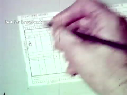 The still depicts a close-up of a hand writing on a form, likely related to educational data management. The context involves IBM's technology from the 1960s, showcasing the use of punch cards and manual data entry to manage grades and course enrollment efficiently. The image suggests a blend of traditional and emerging technology in education during that era.
