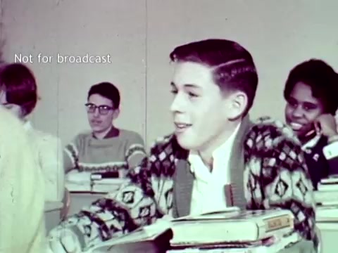 The still features a classroom setting from the 1963 IBM film 'The Information Center.' It shows a group of students seated at desks, with textbooks and materials in front of them. The focus is on a boy wearing a patterned cardigan, smiling as he engages with his classmates, while others appear interested and attentive. This scene likely illustrates the integration of IBM's computing technology in educational contexts, emphasizing the use of tools like punch cards for managing academic tasks such as grades and enrollment.