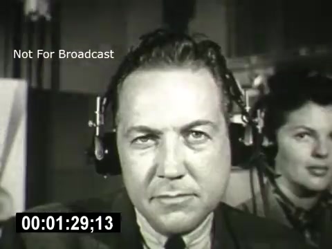 The still features two individuals working in a communication setting, likely a call center or broadcasting environment. The man in the foreground has a serious expression and is wearing headphones, suggesting he is focused on a task. In the background, a woman also wearing headphones appears engaged in the process. The image conveys a sense of professionalism and the importance of communication technology, in line with the themes of the Bell System.