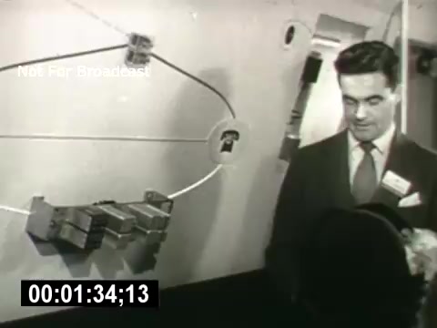 The still from 'Bell System - Story Without End' features a man in a suit, presumably a presenter or spokesperson, interacting with a visual display or model that illustrates telecommunications technology. The background includes a schematic or diagram, possibly representing communication pathways or systems, emphasizing the innovative aspects of the Bell System. The scene captures a moment of explanation or demonstration, reflecting the mid-20th century focus on technological advancement.