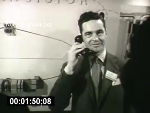 The still depicts a man in a suit, holding a telephone headset to his ear, likely engaged in a conversation. He has a confident smile and is positioned in a technological setting, emphasized by equipment in the background. The image suggests a focus on communication and innovation, aligning with the themes of the Bell System's narrative on technological advancements.