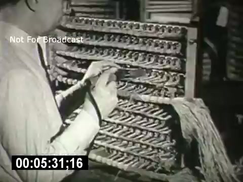 The still features a technician working on a complex network of telephone wires and connections, showcasing the inner workings of telecommunication technology. The focus is on the intricacies of the equipment, highlighting the labor involved in maintaining communication systems. The image appears to be from a historical context, reflecting the era of early telecommunications development.