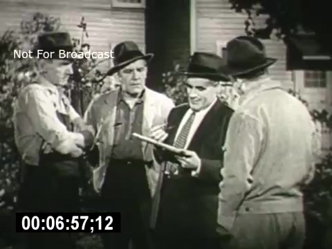 The still from 'Bell System - Story Without End' features four men in hats, gathered outdoors. They appear to be engaged in a discussion, with one man gesturing while holding a clipboard or paper. Their expressions suggest a serious conversation, likely about communication or a local issue. The setting is casual, indicating a community-oriented scenario. The image captures a moment of cooperation and dialogue among the characters.