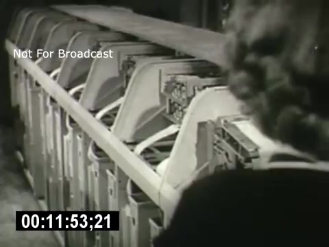 The still from 'Bell System - Story Without End' features a series of machines, likely telecommunications equipment, lined up in a workspace. A person's silhouette is partially visible in the foreground, indicating human interaction with the technology. The image captures a moment of industrial activity, reflecting the era's focus on the expansion and complexity of communication systems. The presence of the timecode suggests that this is a segment from a film, possibly illustrating operational aspects of the Bell System.