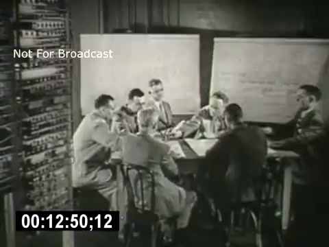 The still from 'Bell System - Story Without End' depicts a group of men gathered around a table, engaged in discussion or analysis. In the background, a series of large boards and a control panel suggest a technical or operational setting, likely related to telecommunications. The focus is on teamwork and collaboration in a professional environment. The black-and-white aesthetic emphasizes the era's style and context.