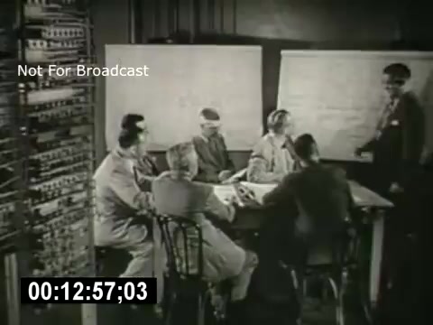 The still features a group of six individuals seated around a table, engaged in discussion. They appear to be in a professional setting, possibly a meeting room. A man is standing and presenting information, while behind him, there are large presentation boards. A piece of machinery or technical equipment is visible on the left, suggesting a focus on telecommunications or technology. The overall tone is formal and collaborative.