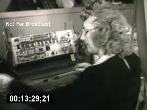 The still from 'Bell System - Story Without End' features a woman with curly hair operating an early telecommunications device. She is engaged in her work, possibly managing calls or connections. The equipment behind her includes an array of dials and switches, indicative of the era's technology. The timecode at the bottom suggests it is part of a larger narrative about the history and evolution of communication.