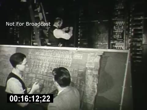 The still from 'Bell System - Story Without End' depicts two men in suits observing a technical panel filled with wires and switches. One man, possibly an operator, is shown working at a control unit, while the second man watches attentively, suggesting a moment of instruction or demonstration. The scene highlights the intricate technology and human interaction involved in telecommunications during that era. The frame is marked with 'Not For Broadcast' and includes a timecode at the bottom.