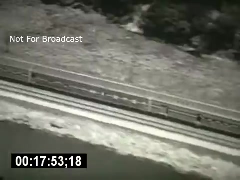 The still depicts a black-and-white scene featuring a road or railway track alongside a body of water or riverbank. The image has a timecode overlay at the bottom, indicating a specific moment in the film. The overall composition suggests a focus on transportation or communication infrastructure, aligning with the themes of the Bell System. The context implies a documentary style, highlighting the interconnectedness of technology and the environment.