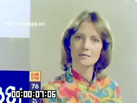 The still features a woman with shoulder-length hair, wearing a colorful, floral-patterned blouse. She appears to be engaged and looking directly at the camera, possibly conveying a message or information. The background includes a display with Kodak branding and a timestamp in the lower corner. The overall aesthetic reflects the vibrant style characteristic of the late 1970s.