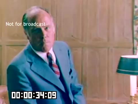 The still features a middle-aged man in a tailored blue suit with a red and white striped tie, sitting against a wooden paneled wall. He appears to be looking directly at the camera with a serious expression. A lamp is visible on a table beside him. The image contains a 'Not for broadcast' label and a timestamp. The context suggests it’s part of a promotional film for Kodak, featuring notable personalities from the 1970s.