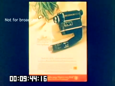 The still features a promotional advertisement for a Kodak camera from 1977, showcased prominently against a colorful background. The camera, likely a model designed for filmmaking, is displayed in a sleek, modern design typical of that era. Text on the ad highlights features or benefits of the camera, while a 'Not for broadcast' label indicates its internal use. The timestamp at the bottom suggests this is part of a longer sequence.