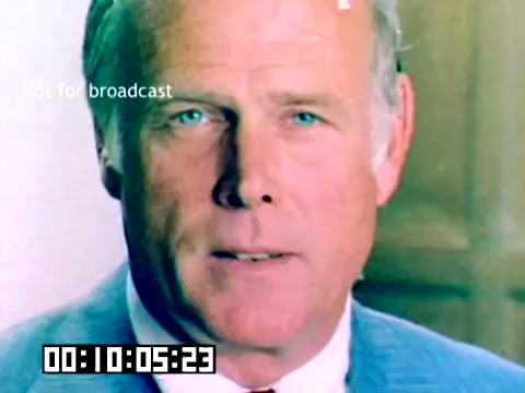 The still features a close-up of a man with blue eyes and light hair, dressed in a blue suit. He appears to be speaking or presenting, showcasing a focused expression. The background is a soft focus, suggesting a formal setting. There is a timecode visible at the bottom and a watermark stating 'Not for broadcast.' The image is from a silent Kodak sales film, likely featuring personalities associated with the brand in 1977.