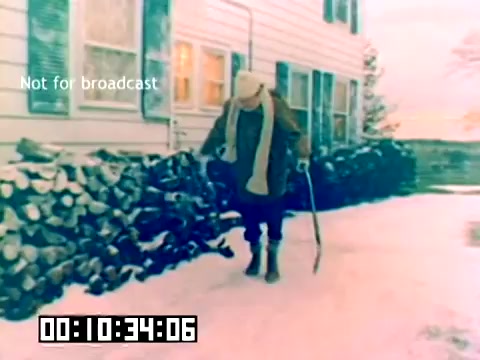 The still features a snowy outdoor scene with a figure walking along a path next to a stacked pile of firewood. The figure, dressed warmly with a scarf and hat, uses a cane while navigating through the snow. A house is visible in the background, with a few windows that suggest a cozy, residential setting. The timestamp on the bottom indicates it is from 10 minutes and 34 seconds into the footage, and 'Not for broadcast' is noted on the image. The overall atmosphere conveys a wintery, peaceful moment.