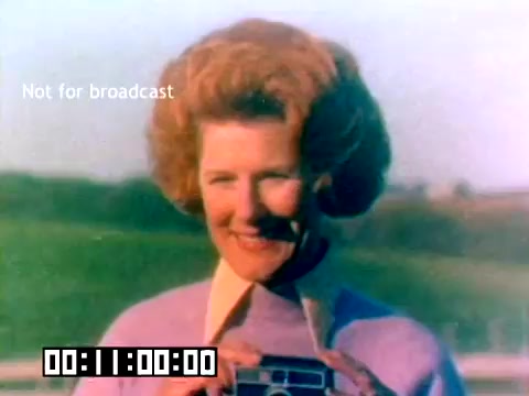 The still captures a smiling woman with voluminous, curly hair, dressed in a pastel purple sweater. She holds a vintage Kodak camera and appears to be outdoors, with a green landscape in the background. The image is marked 'Not for broadcast' and features a timestamp at the bottom. The quality reflects the aesthetics of 1970s film. This promotional still likely aimed to convey the joy of photography and the use of Kodak products.