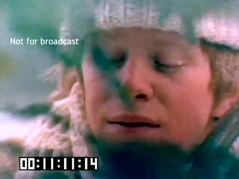 The still from the 1977 Kodak sales film features a close-up of a young man with a thoughtful expression, dressed warmly in a knitted hat and scarf. The image is tinged with a soft focus, suggesting a nostalgic or intimate moment. In the background, there are hints of a wintery landscape, adding to the serene atmosphere. A watermark reads 'Not for broadcast,' indicating the film's private or promotional nature. The time code displays the elapsed time, enhancing the film's vintage quality.