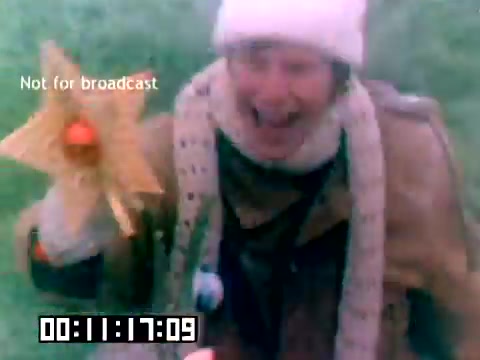 The still from the 1977 Kodak sales film features a man enthusiastically holding a paper flower with an orange center, wearing a winter hat and a scarf. He appears to be laughing or shouting joyfully, set against a grassy background. The image includes a timestamp and a 'Not for broadcast' label, indicating its promotional nature. The vibrant colors and the man's expressive demeanor suggest a lighthearted context, typical of sales films aimed at showcasing Kodak products in a fun, engaging way.