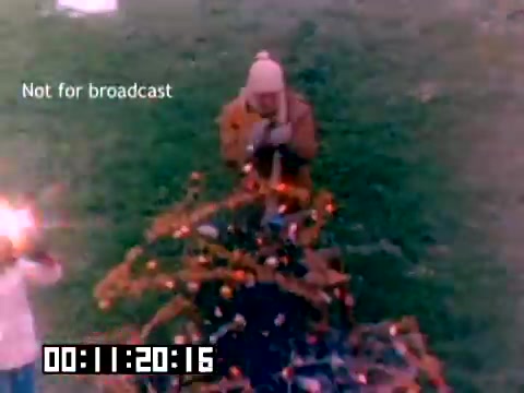 The still features a scene from a 1977 Kodak sales film, showing a person in a beanie and orange jacket engaging with a Christmas tree adorned with lights. Another figure, possibly a child, is nearby, holding sparklers. The image is slightly faded, characteristic of older film stock, and includes a watermark stating 'Not for broadcast.' A timecode appears at the bottom, indicating the moment in the footage. The lack of sound adds an eerie quality to the nostalgic visuals.