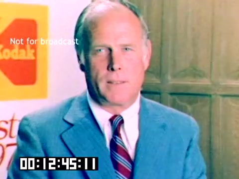 The still features a middle-aged man in a light blue suit with a striped tie, standing in front of a background displaying the Kodak logo. The setting appears professional, likely from a promotional or sales presentation. The timecode at the bottom indicates a specific moment in the footage, and there's a watermark stating 'Not for broadcast,' suggesting it's an internal or restricted viewing material. The overall atmosphere is formal, reflecting a marketing context from 1977.
