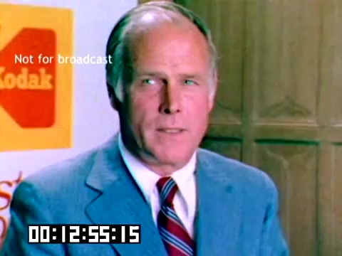 The still showcases a middle-aged man with a confident demeanor, likely a presenter or spokesperson at a Kodak event in 1977. He is dressed in a suit and tie, with a backdrop featuring the Kodak logo prominently displayed. The image is colorful and conveys a professional setting, typical of sales presentations from that era. The time code and 'Not for broadcast' watermark suggest this is a production still rather than a public release.
