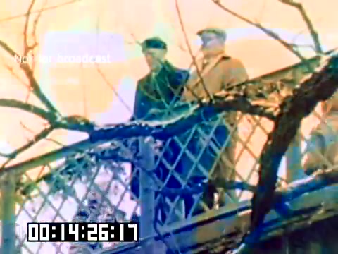 The still captures two figures standing on a balcony or walkway, framed by a lattice structure. The men appear to be dressed in coats and hats, likely reflecting the fashion of the late 1970s. The background features a bright, colorful backdrop, possibly a sky or set design, creating a vivid contrast against the muted tones of their clothing. The overall mood suggests a moment of reflection or observation in this silent Kodak sales film.