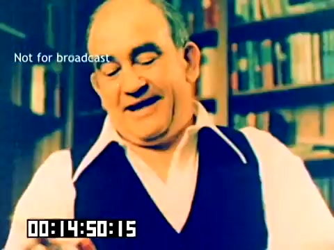 The still is a colorful, 16mm image featuring a man, likely presenting or demonstrating a Kodak product. He is dressed in a collared shirt and a vest, smiling as he interacts with the camera. In the background, there are bookshelves filled with books, suggesting a cozy setting. The text 'Not for broadcast' and a time stamp appear at the bottom of the frame, indicating that it's a sales film from 1977. The absence of sound leaves the viewer to interpret his expression and gestures without audio context.