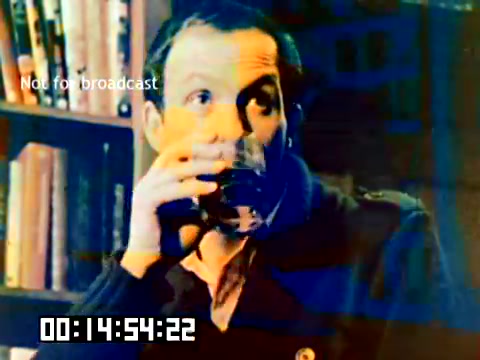 The still features a man holding a glass, likely engaged in conversation or reflection. He is dressed in casual attire, and the background includes shelves of books, suggesting a cozy indoor setting. The image has a slightly distressed quality, indicating its age from the 1977 Kodak sales film. The timestamp in the corner indicates it's part of a larger sequence. The muted colors and focus on the subject convey a sense of nostalgia.