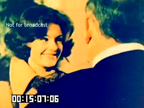 The still features a woman smiling warmly, engaging with another individual who is partially visible, suggesting a friendly or affectionate interaction. The colors appear vibrant, indicative of the period's film quality. Overlay text indicates that it is not for broadcast, and a timecode is present at the bottom. The atmosphere seems light and positive, reflecting the promotional nature of the Kodak sales film from 1977.