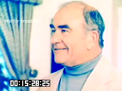 The still features a smiling older man, dressed in a light-colored suit with a turtleneck. He has a friendly expression, conveying a sense of warmth and engagement. The background includes soft furnishings, hinting at a domestic or professional setting. The image is part of a silent Kodak sales film from 1977, featuring notable personalities like Dick Van Dyke and Michael Landon, although their presence is not depicted in this particular frame. The lower corner displays a timecode and a note indicating the film is not for broadcast.
