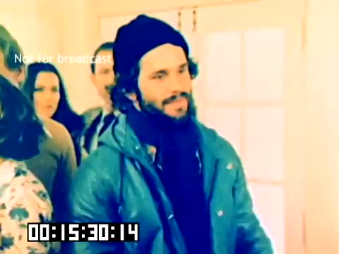 The still appears to show a scene from a 1977 Kodak sales film featuring a group of individuals. A man with curly hair and a beard, wearing a blue jacket and black beanie, is in the foreground, looking somewhat engaged or contemplative. He is surrounded by a few other people, with a woman in a patterned outfit visible in the background. The overall aesthetic is colorful, indicative of the 70s era, although the lack of sound makes it difficult to discern the full context of the scene. The timestamp at the bottom indicates it is 15 minutes and 30 seconds into the footage.