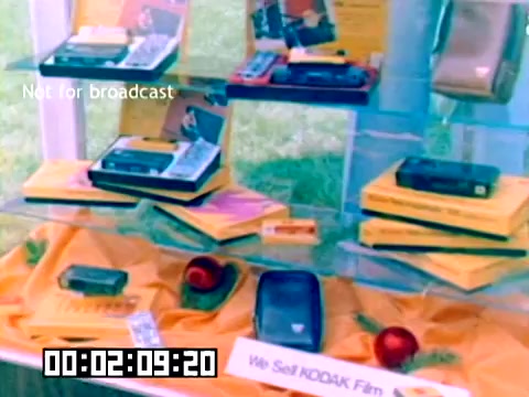 The still captures a display showcasing Kodak products from 1977. Prominently featured are various cameras and film packages arranged on an orange tablecloth. A sign reading 'We Sell KODAK Film' highlights the focus on Kodak’s film products. The overall presentation reflects the marketing style of the era, emphasizing the technology and consumer goods offered by Kodak.