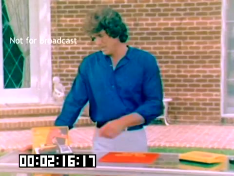 The still features a young man with curly hair, wearing a blue shirt, standing at a table with colorful Kodak products, including boxes and possibly cameras. The background shows a brick wall and a window, suggesting an outdoor setting. The timestamp indicates 2 minutes and 16 seconds into the film, and a 'Not for broadcast' label is visible. The overall tone reflects the marketing style of the late 1970s, aimed at promoting Kodak's photography products.