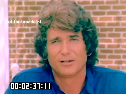 The still features Michael Landon, sporting a hairstyle typical of the 1970s, with medium-length, wavy hair. He is wearing a blue shirt and is positioned against a backdrop of brickwork. The image includes a timestamp and a note indicating that it is 'not for broadcast.' The overall aesthetic reflects the era's color grading and style.