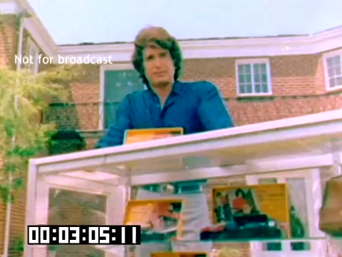 The still captures a key moment from a 1977 Kodak sales film. A man, dressed in a blue shirt, stands in front of a display case filled with Kodak products, likely showcasing film or cameras. The backdrop features a brick building and greenery, indicating an outdoor setting. The timestamp and 'Not for broadcast' label suggest it's an excerpt meant for internal or promotional use. The overall tone is promotional, aiming to attract interest in Kodak's photography products.