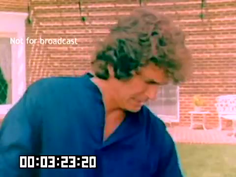 The still shows a man with curly hair wearing a blue shirt, outdoors in a backyard setting. The background features a brick wall and some garden furniture. There’s a timestamp at the bottom indicating the film's progression. The frame is marked as 'Not for broadcast,' suggesting it was intended for internal use only. This snapshot captures a casual moment from a Kodak sales film featuring notable figures like Dick Van Dyke and Michael Landon from 1977.