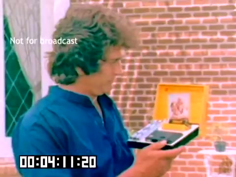 The still features a man with curly hair, holding a Kodak product case while standing in front of a brick wall. He is dressed in a blue shirt and appears to be demonstrating the item, which includes a box that opens to reveal a camera and accessories. The caption 'Not for broadcast' indicates it's a behind-the-scenes or promotional piece. The timestamp at the bottom suggests the scene is part of a longer film. The overall setting has a casual, home-like atmosphere, typical of sales presentations from the late 1970s.