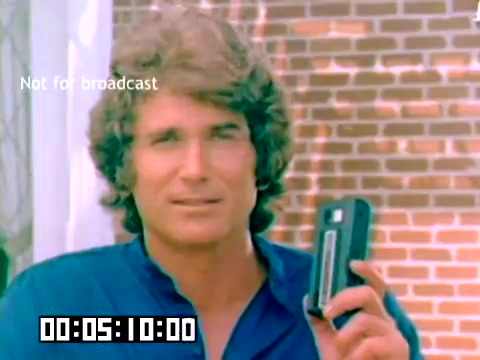 The still features a young Michael Landon, wearing a blue shirt, and holding a compact Kodak camera. He has slightly wavy hair and a friendly expression. The background shows a brick wall, suggesting an outdoor setting. A digital timer indicates the moment in the film, and a label notes that the image is 'Not for broadcast.' The still captures a promotional tone typical of 1970s sales films, focusing on stylish, user-friendly photography equipment.