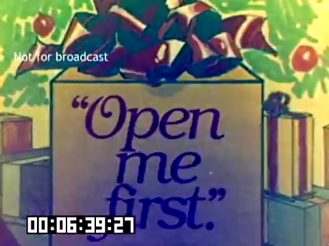 The still features a festive gift box adorned with a large bow, set against a colorful, holiday-themed background. The text 'Open me first.' is prominently displayed on the side of the box, suggesting a message of importance or priority. The overall tone is cheerful and inviting, typical of holiday marketing. The scene likely aims to capture attention and promote a product, in line with Kodak's sales strategies in 1977.