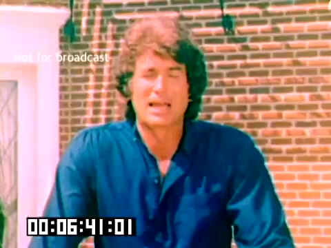The still features a man with long, wavy hair, wearing a blue shirt, standing in front of a brick wall. He appears to be speaking or presenting to the camera, conveying enthusiasm or engaging with the audience. The image includes a timestamp at the bottom and a watermark indicating it is not for broadcast. The overall style reflects the aesthetics of 1970s promotional films.
