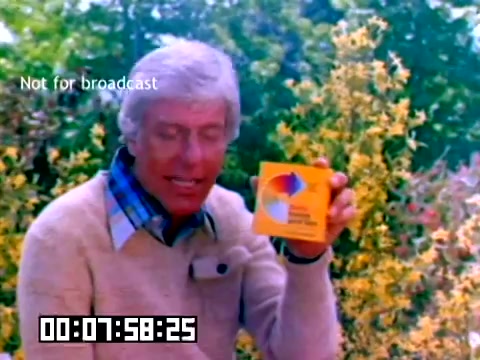 The still from the silent Kodak sales film features a man with silver hair, dressed in a light sweater over a collared shirt, presenting a box of Kodak film. The background is filled with vibrant yellow flowers, suggesting a bright and cheerful outdoor setting. The overlay indicates 'Not for broadcast,' and a timestamp appears in the lower left corner. The context revolves around a promotional piece from 1977, highlighting Kodak's products, though the absence of audio limits the understanding of the presentation.